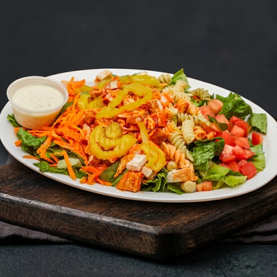 A colorful salad with chicken, pasta, shredded carrots, tomatoes, and banana peppers, with a side of dressing.