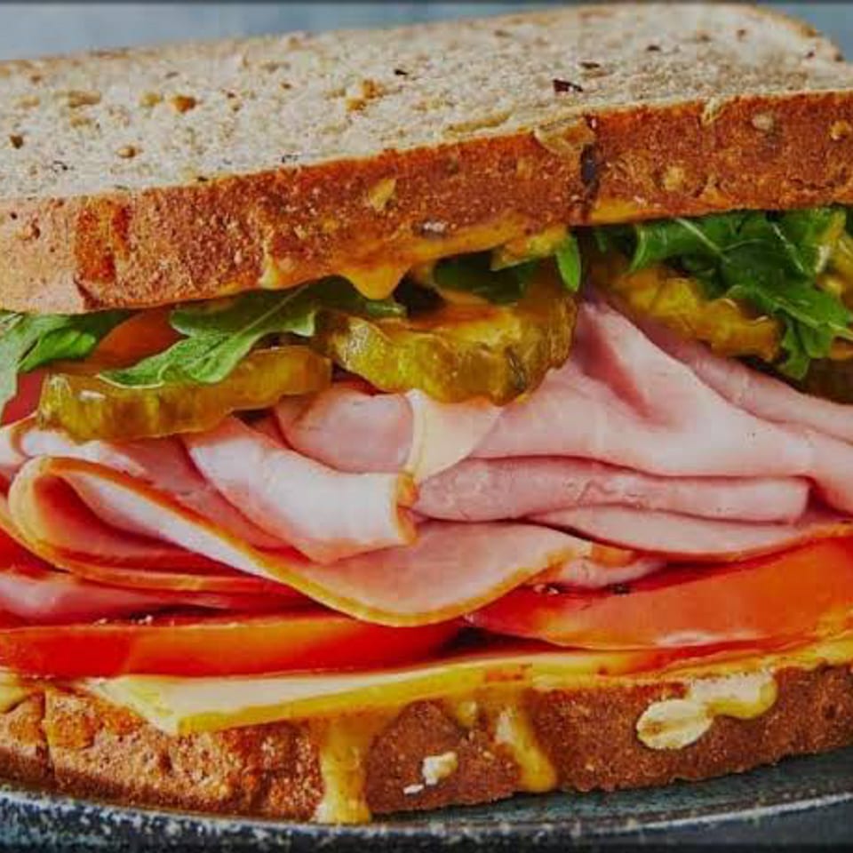 Overstuffed toasted deli sandwich with ham, melted cheese, tomato, pickles, and leafy greens on a dark plate.