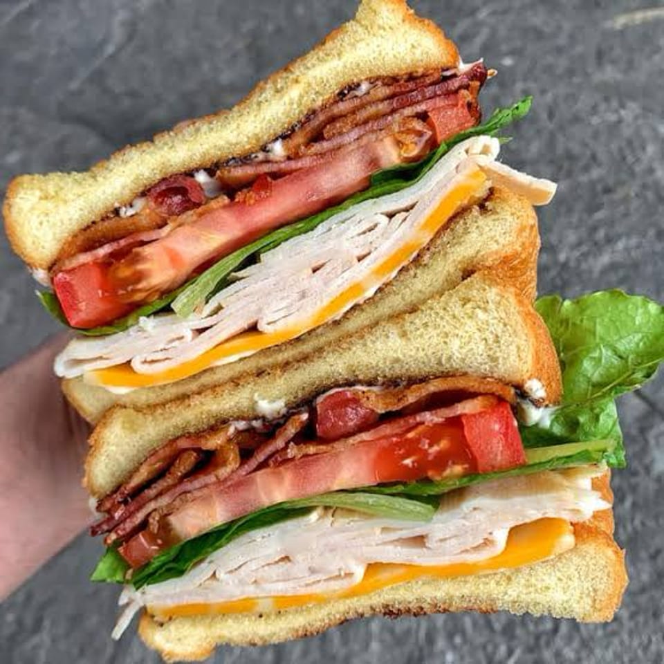 Stacked club sandwich halves with turkey, bacon, ham, cheese, lettuce, and tomato on toasted bread.