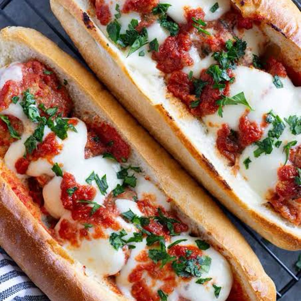 Two meatball subs with melted mozzarella, marinara sauce, and fresh parsley on a cooling rack.