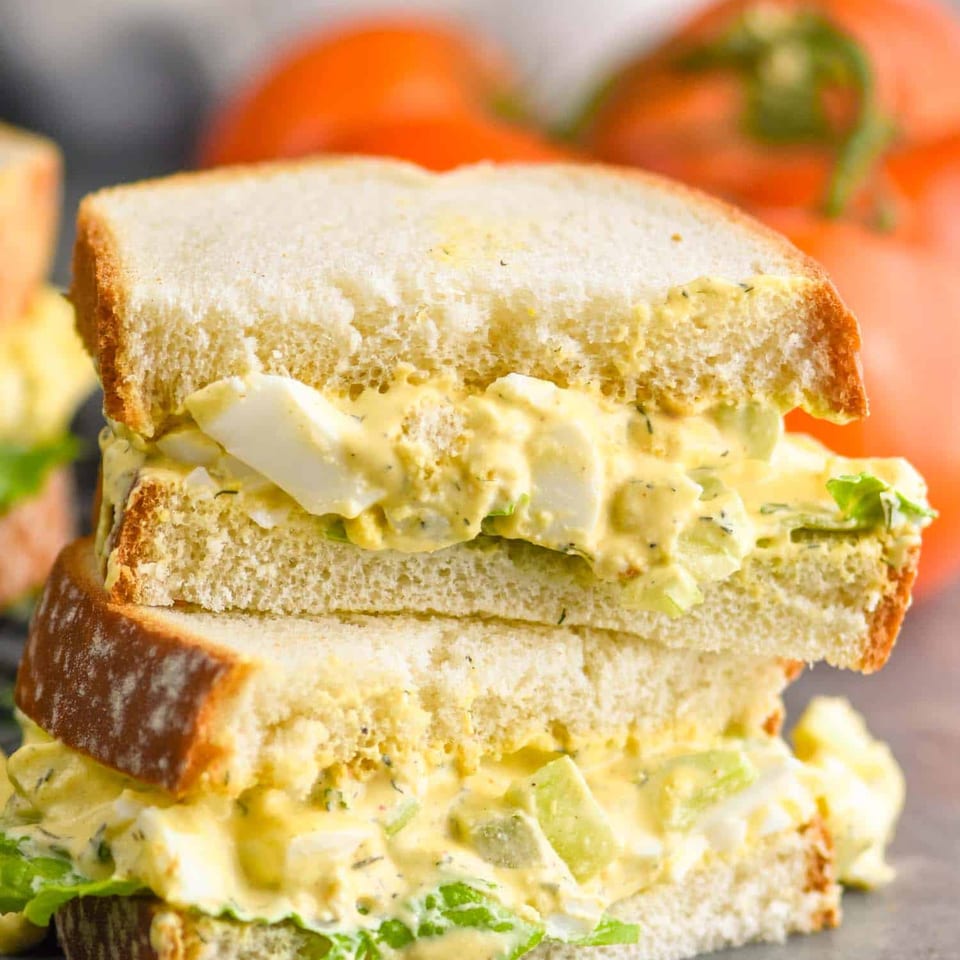 Stacked egg salad sandwiches with creamy herb filling, lettuce, and tomatoes in the background.