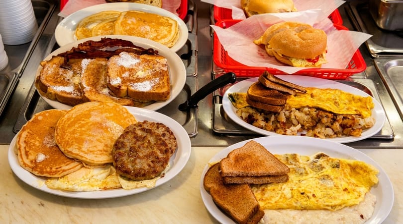 Multiple plates of breakfast food, including pancakes, French toast, bacon, sausage, omelets, and hash browns.