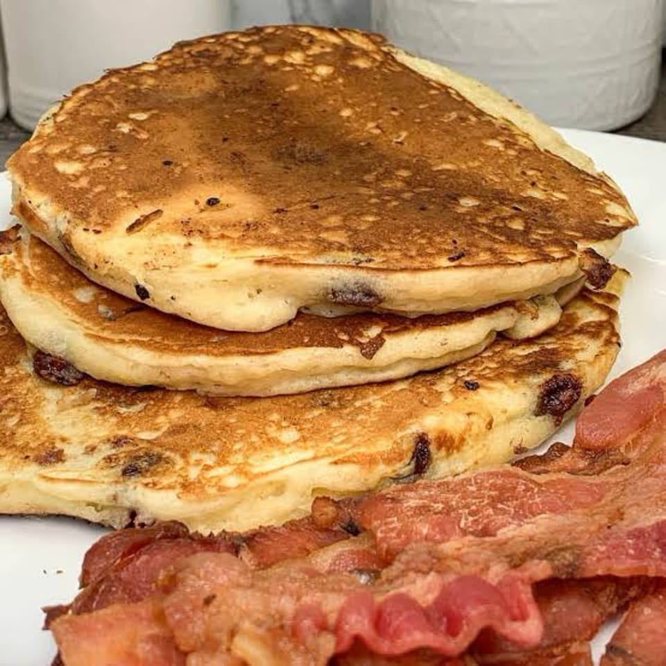 Stack of golden-brown pancakes with chocolate chips and crispy bacon on a white plate.